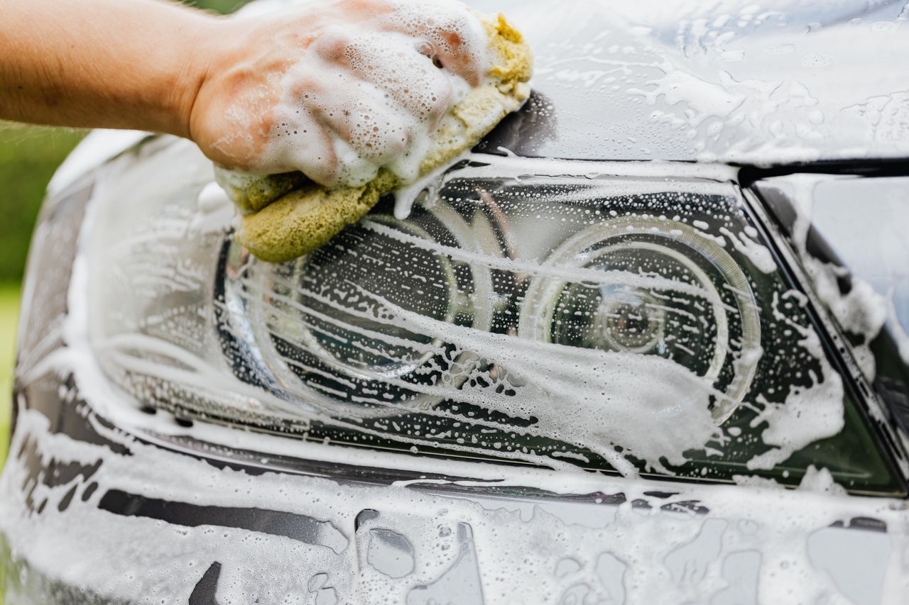 Hand scrubbing a car headlight with sponge and soap suds, detailing process.