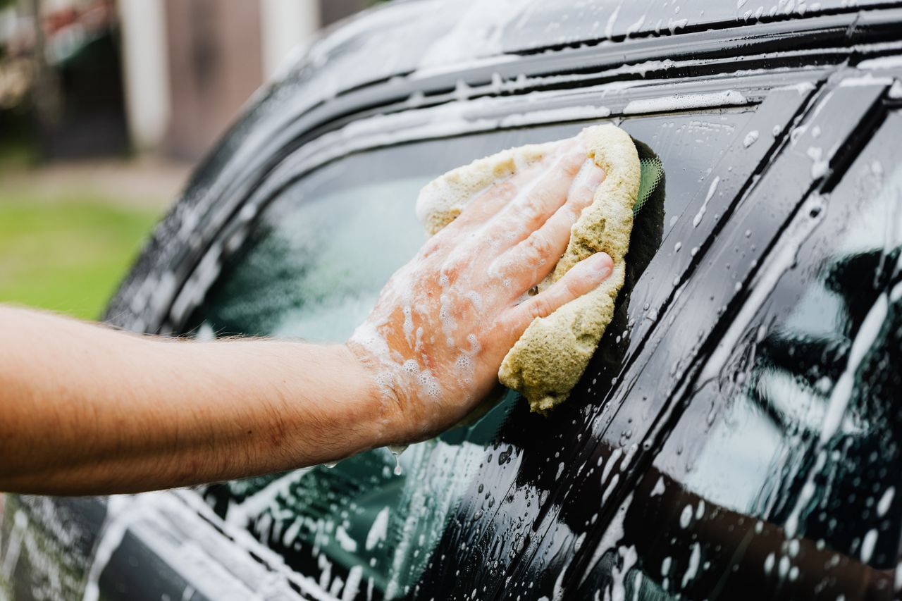 About A hand scrubs a car window with a sponge, covered in soap suds, during a car wash.